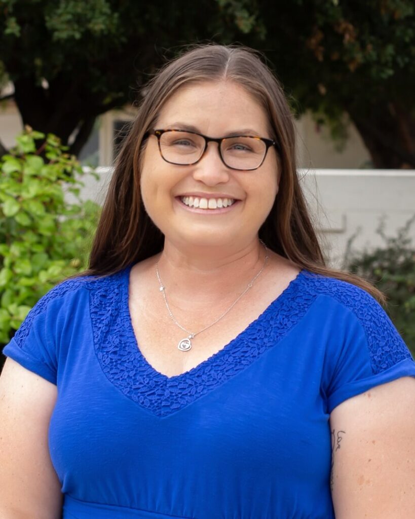 A person with long brown hair smiling wearing black glasses, necklace, and blue shirt.