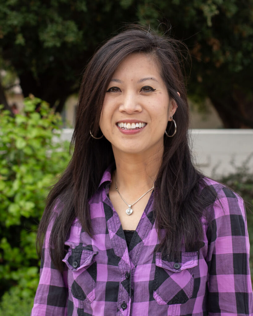 A person with long black hair, necklace, wearing a purple plaid shirt.