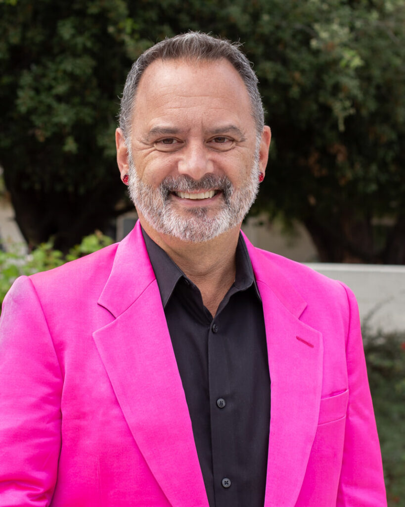 A person with short hair smiling wearing a pink coat and black shirt.